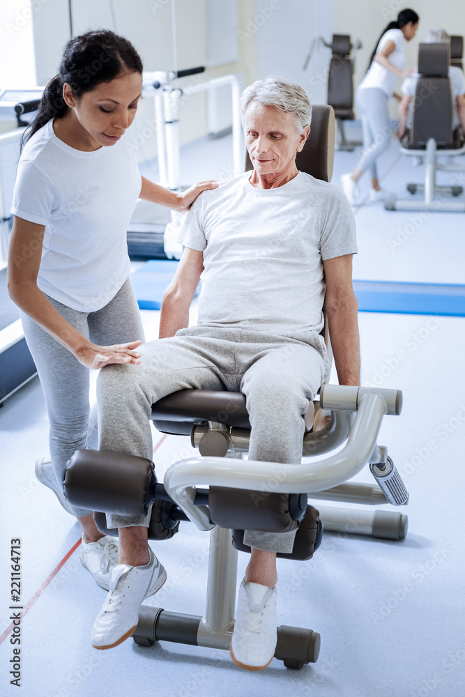 Very careful. Calm serious senior man looking attentive while sitting on a special exercise machine with a qualified reliable specialist of a rehabilitation center examining his injured knee Very careful. Calm serious senior man looking attentive while sitting on a special exercise machine with a qualified reliable specialist of a rehabilitation center examining his injured knee