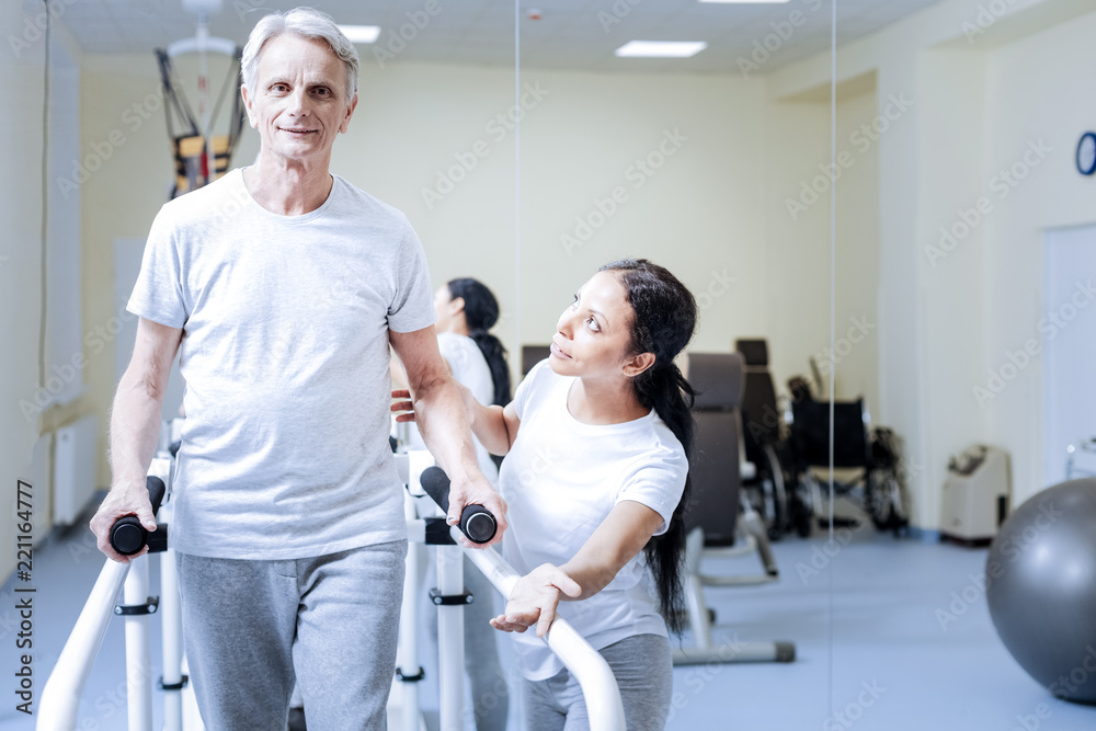 Try it. Polite kind experienced medical worker encouraging her aged disabled patient to make a few steps on a convenient treadmill