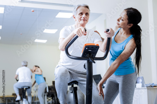 Nice activity. Kind attentive qualified trainer looking at her energetic recovering patient on an exercise bike and smiling while noticing his progress