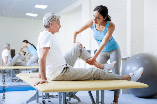 Stretching. Attentive responsible calm medical worker being careful while warming the injured muscles of a worried aged patient in the rehabilitation center