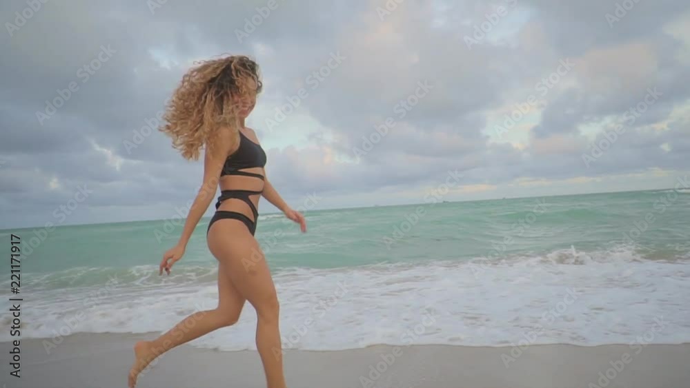 Beautiful girl is fleeing on sandy beach of ocean in summer day. She is smiling and looking at camera, in background of cloudy sky Beautiful girl is fleeing on sandy beach of ocean in summer day. She is smiling and looking at camera, in background of cloudy sky