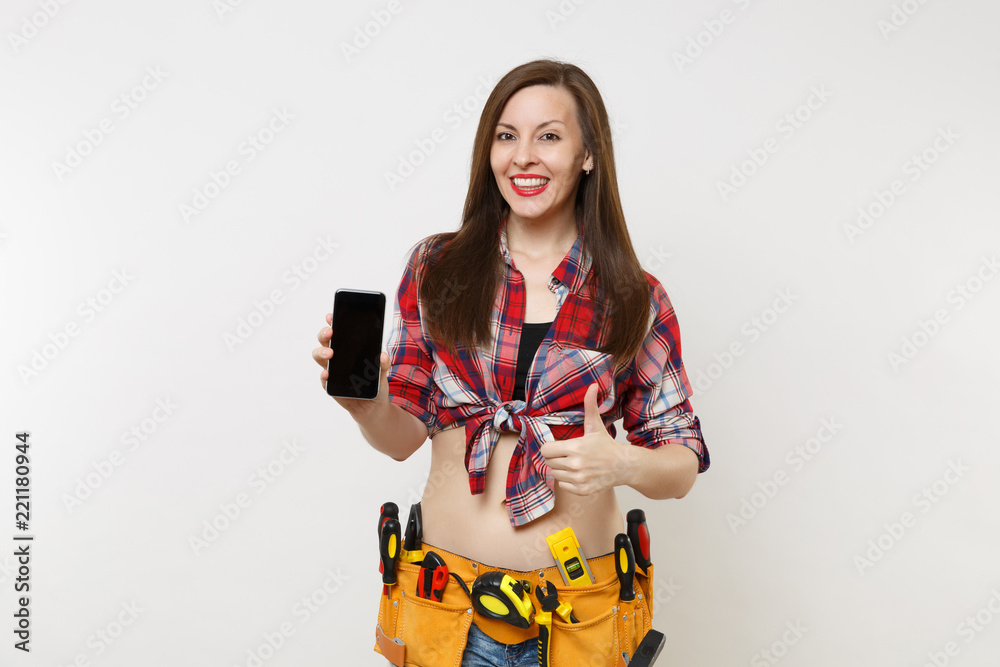 Woman in kit tools belt full of instruments holding showing camera mobile phone with blank black empty screen display touchscreen isolated on white background. Female in male work. Renovation concept. Woman in kit tools belt full of instruments holding showing camera mobile phone with blank black empty screen display touchscreen isolated on white background. Female in male work. Renovation concept.