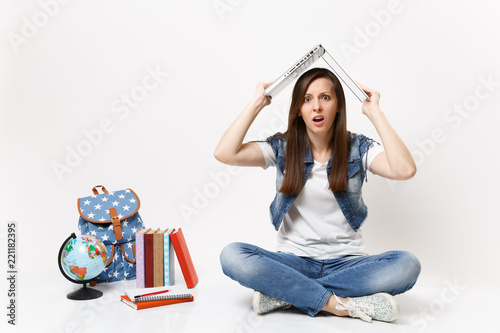 Young exhausted bewildered woman student holding laptop pc computer above head like roof sit near globe backpack school books isolated on white background. Education in high school university college.