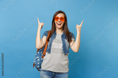 Young shocked amazed woman student with opened mouth with backpack in orange heart glasses pointing index fingers up isolated on blue background. Education in university. Copy space for advertisement.