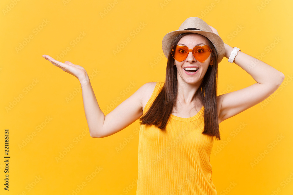 Portrait of smiling young woman in straw summer hat, orange glasses pointing right hand aside copy space isolated on yellow background. People sincere emotions, lifestyle concept. Advertising area.