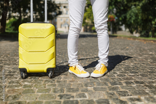 Cropped image of fun traveler tourist woman legs in yellow summer casual clothes with suitcase on road in city outdoor. Girl traveling abroad to travel on weekends getaway. Tourism journey lifestyle.