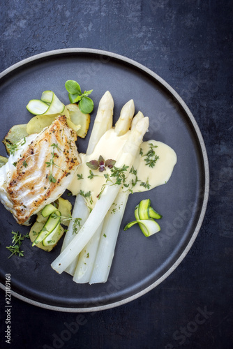Modern Style German fried cod fish filet with white asparagus in hollandaise sauce with roast potatoes and sliced zucchini as top view on a plate with copy space