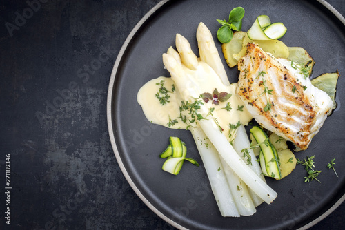 Modern German fried cod fish filet with white asparagus in hollandaise sauce with roast potatoes and sliced zucchini as top view on a plate with copy space left