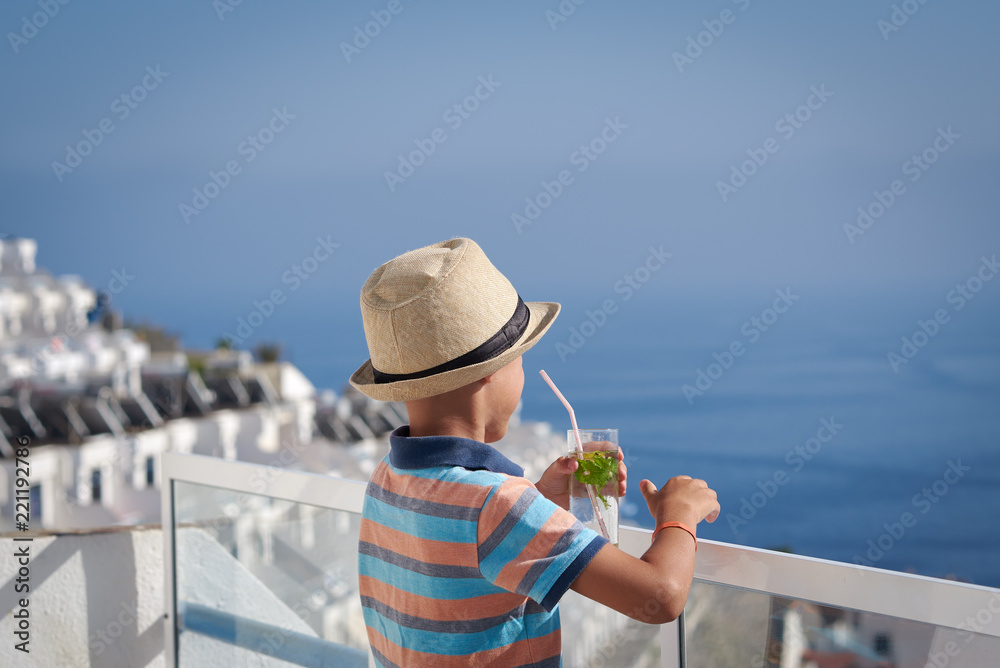 Cute european boy is standing on the hotel's balcony in front of the ocean and holding cocktail in his arm.