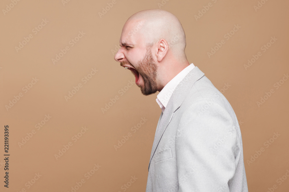 Profile side view portrait crazy anger of middle aged bald bearded businessman in light gray suit standing and screaming with oepn mouth. indoor studio shot, isolated on light brown background. Profile side view portrait crazy anger of middle aged bald bearded businessman in light gray suit standing and screaming with oepn mouth. indoor studio shot, isolated on light brown background.