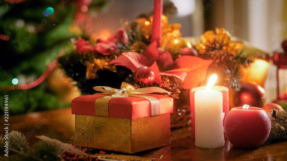 Closeup image of gift box with golden ribbon and burning candles against Christmas tree at living room
