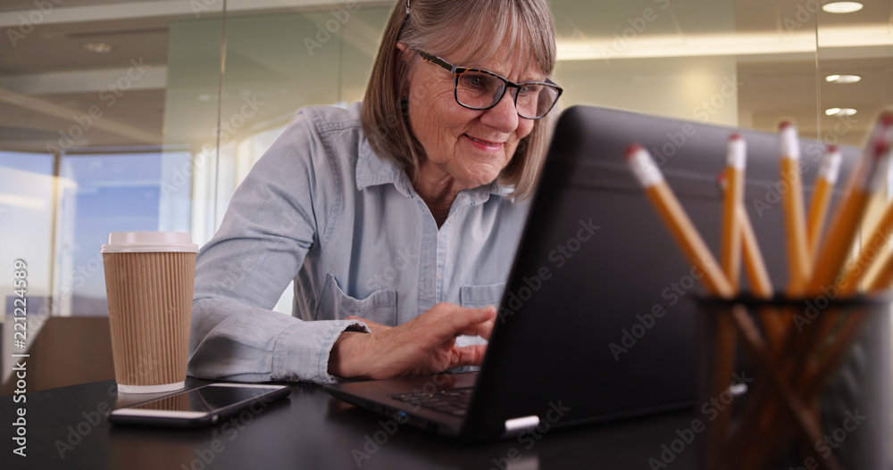 Happy senior woman using laptop inside in modern office setting Happy senior woman using laptop inside in modern office setting