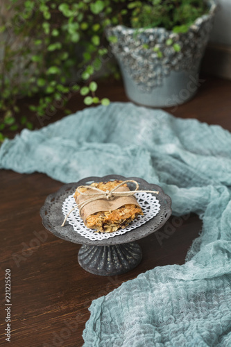 Homemade Chewy Granola Bars on a Tin Pedestal on a Wood Table; Granola Bar Wrapped in Parchment Paper and String; White Heart-Shaped Doily on Pedestal photo