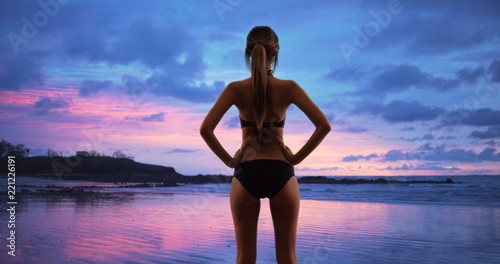 Rear view of fit woman in black swimsuit with hands on hips at serene beach