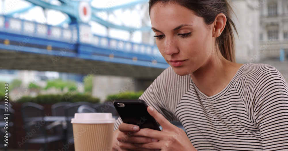 Young Caucasian lady with coffee cup messaging on phone seated by Tower Bridge Young Caucasian lady with coffee cup messaging on phone seated by Tower Bridge