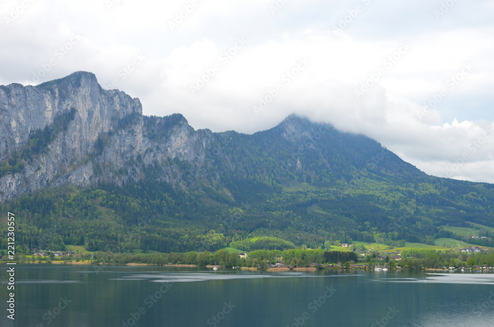 A mountain dwarfs a small village which is on the edge of a lake. Storm clouds fill the sky. The lower slopes of the mountain are covered in forest. A mountain dwarfs a small village which is on the edge of a lake. Storm clouds fill the sky. The lower slopes of the mountain are covered in forest.