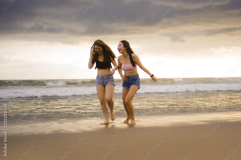 two happy and attractive young Asian Chinese women girlfriends or sisters having fun walking on the beach laughing and talking beautiful light enjoying summer holidays