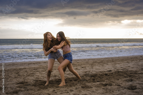 two happy and attractive young Asian Chinese women girlfriends or sisters having fun playing wrestling on sunset beach in beautiful light enjoying summer holidays