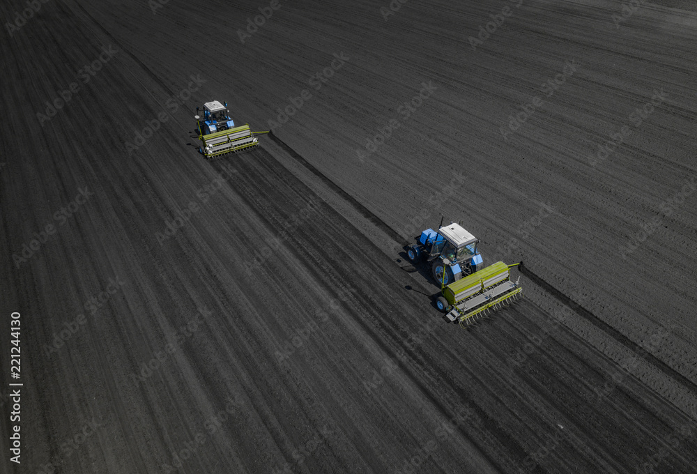Fototapeta premium Aerial view of two blue tractors plows the earth in field on a summer day against a black earth background. Agriculture. Two tractors travel one after another along the black field