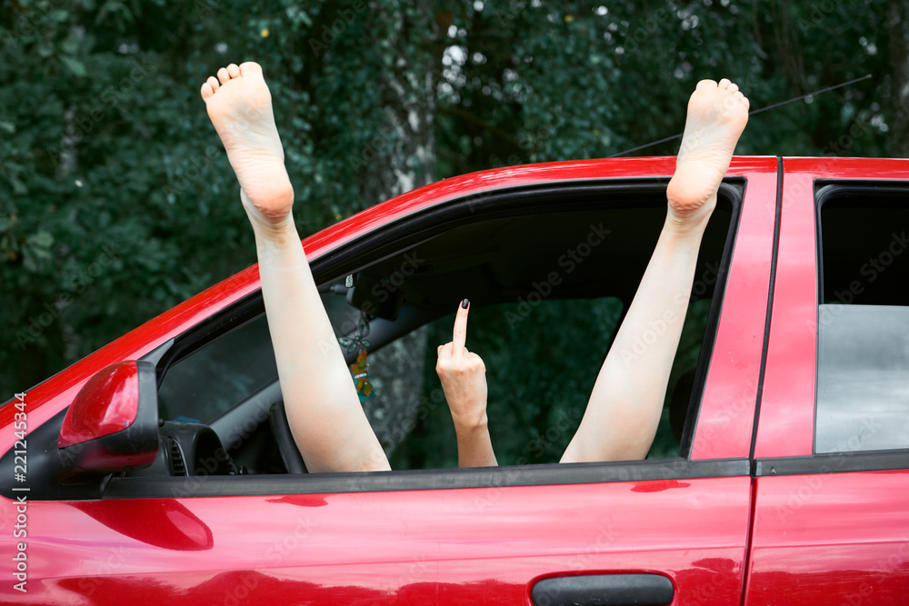 young woman driver resting in a red car, put her feet on the car window and  shows the indecent gesture with middle finger Photos