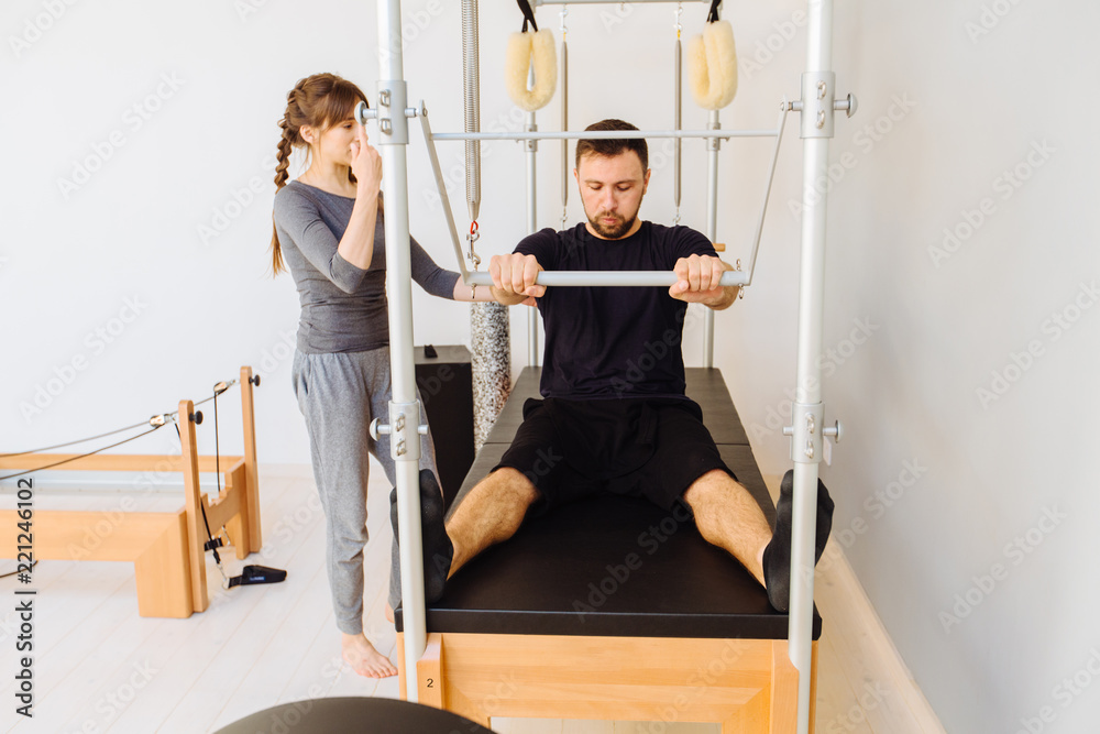 Young sporty female instructor of pilates helping beard man workout in cadillac bed. Two people working in pilates studio, woman assistant supporting and correcting male patient beginner.