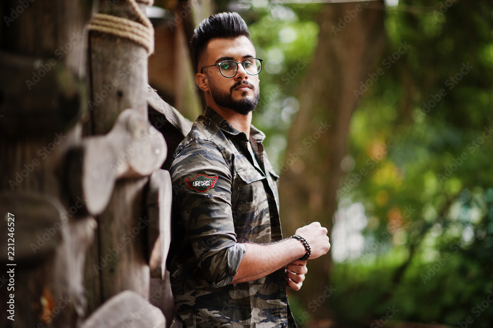Awesome beautiful tall ararbian beard macho man in glasses and military jacket posed outdoor against wooden rustic house.