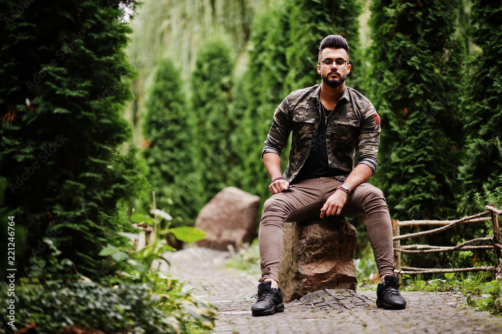 Awesome beautiful tall ararbian beard macho man in glasses and military jacket posed outdoor, sitting on stone.