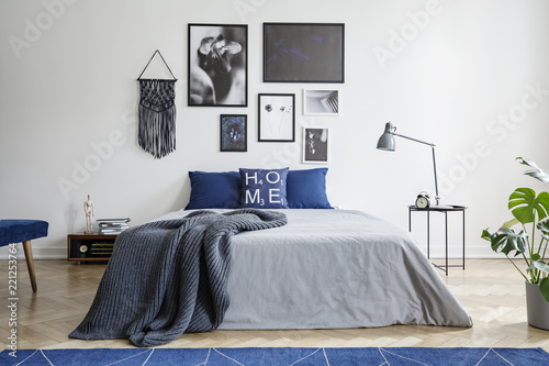 Blanket on bed with blue pillows in white bedroom interior with gallery and lamp on table. Real photo