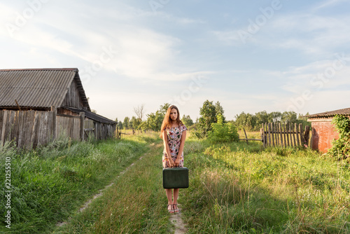 a model girl in a retro dress is holding in her hands a vintage suitcase on an abandoned country road overgrown with grass between the village courtyards and huts