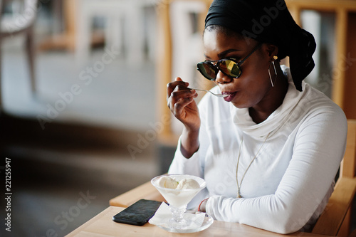 African muslim girl in black hijab and sunglasses sitting at cafe and eating ice cream.
