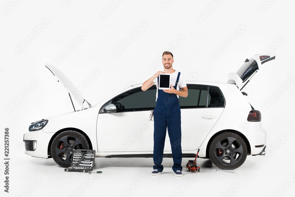 smiling auto mechanic showing tablet with blank screen near broken car on white