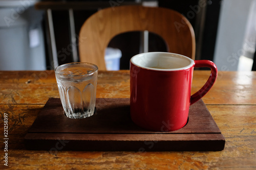 Red cup of coffee and a glass of iced water on wooden tray on rustic wooden table and wooden chair in background in cafe