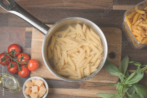 Cooking pasta and ingredients on a wooden table with cutting board, dark background, top view. Italian food concept. Pasta penne, tomatoes, basil, parmesan, flat lay