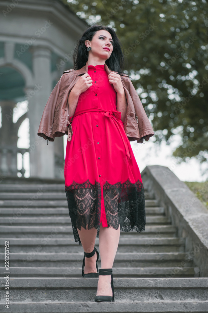 Attractive young woman in red dress and brown leather jacket  walking down the stairs in the park