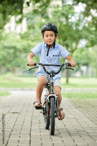 Cheerful little Asian boy in helmet riding bicycle in park