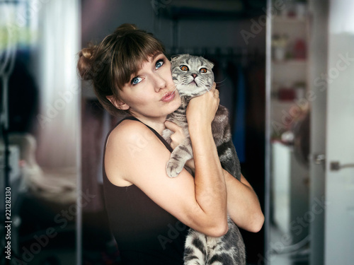 Beautiful young blonde woman in black t-shirt with high buns hairstyle looking with easy attractive smile petting scottish fold cat indoors in her room photo