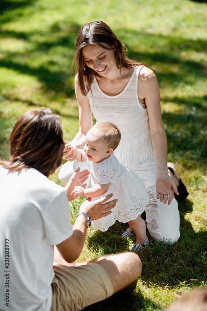 Fototapeta premium Smiling parents dressed in the white clothes are teaching their little daughter how to make her first steps on the lawn.