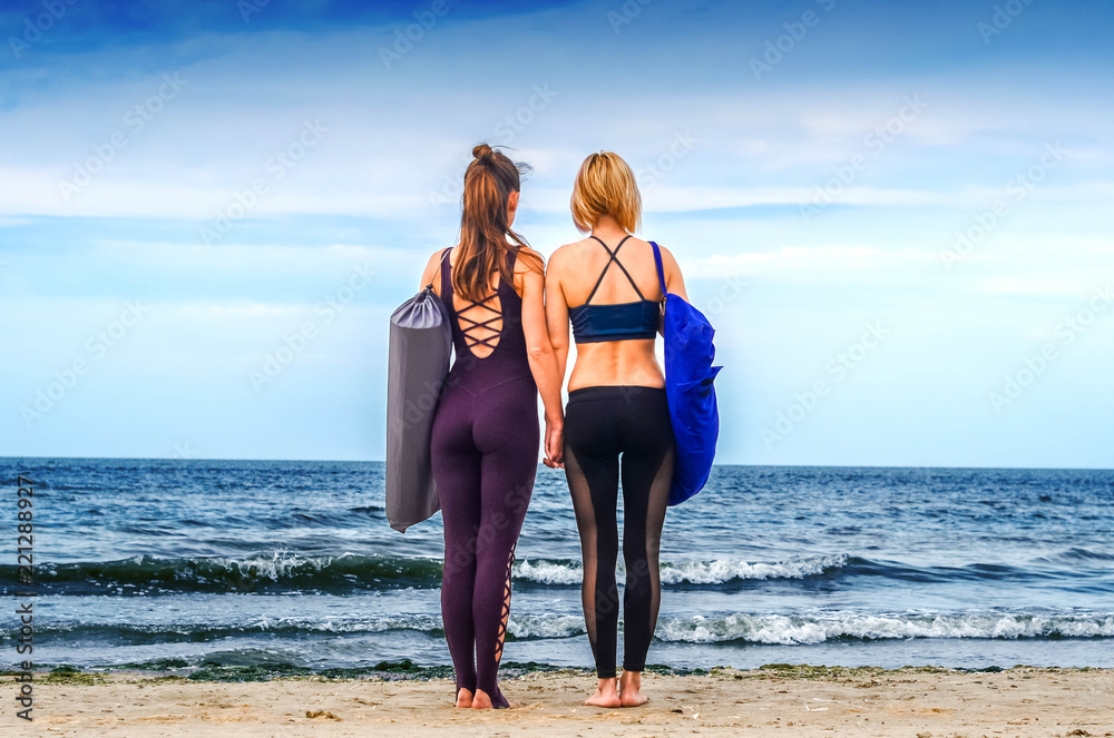 two girls with yoga mats in their covers hold hands and look forward on the horizon on the shore of the blue sea on a clear day