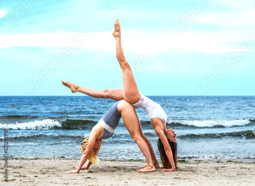 two young girls in white top and black bottom on the sandy shore of the blue sea stand in a pair yoga asana on a clear day