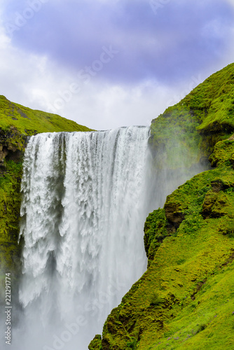 Beautiful Iceland Waterfall.Fast running river. Rugged volcanic rock cliffs against blue skies with low clouds. Green moss and grass covered rock formation.