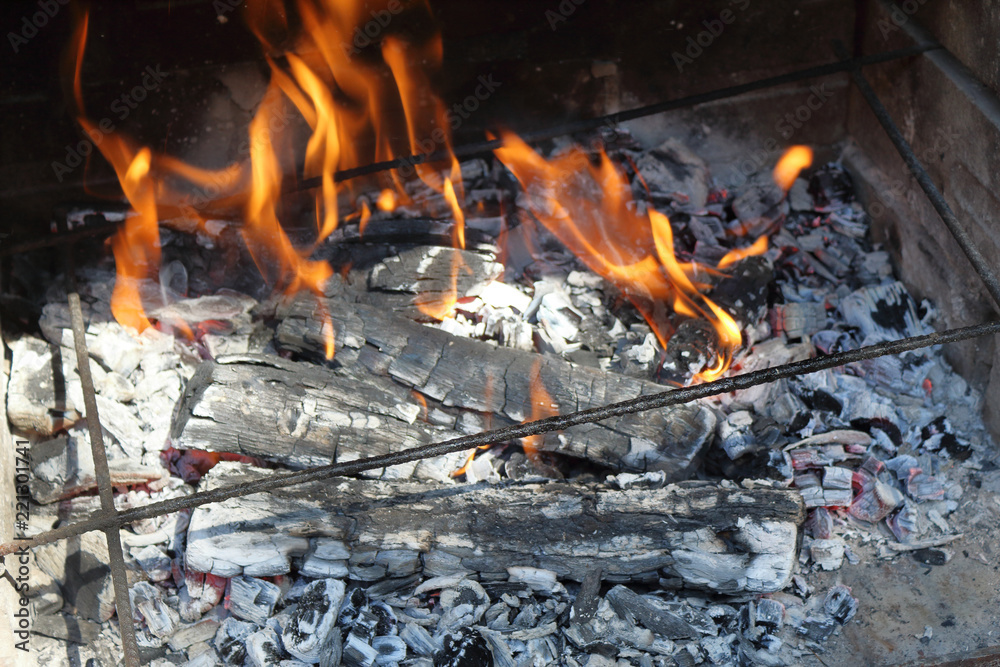 A close photo of firewood burning for a barbecue in a sunny day, with ash, carbon and flames