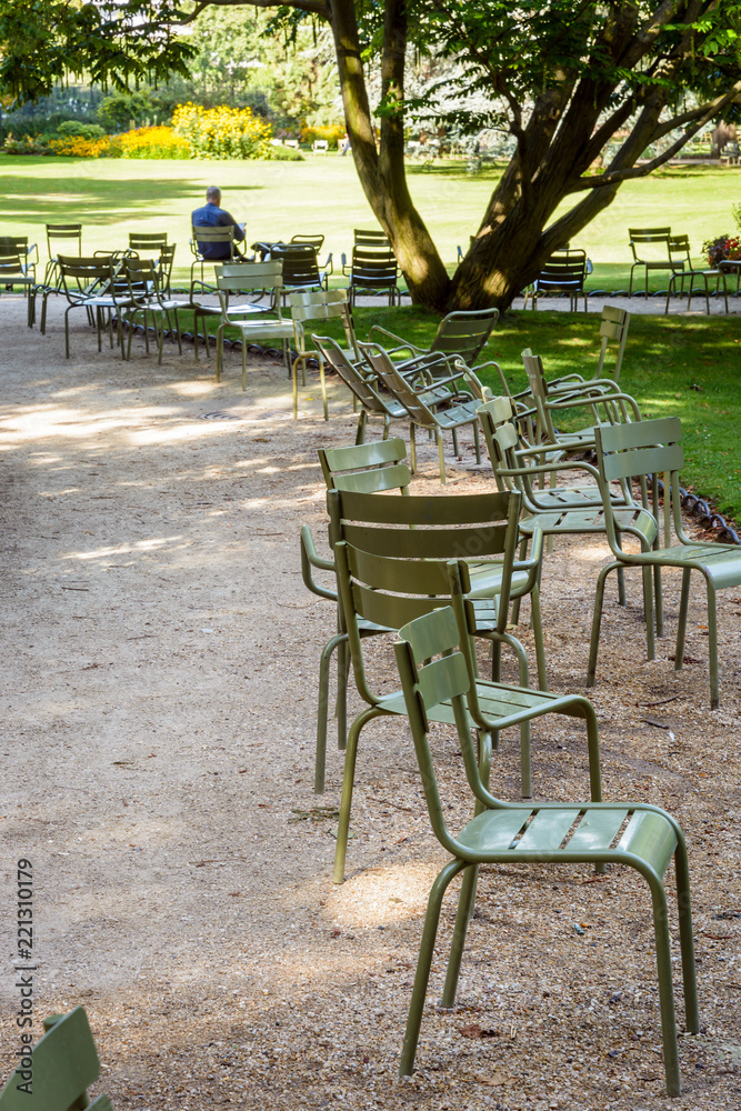 A man alone reading a book, seated in the shade on a metal lawn chair in the Luxembourg garden in Paris, France, by a sunny morning with seats scattered alongside the alleys.