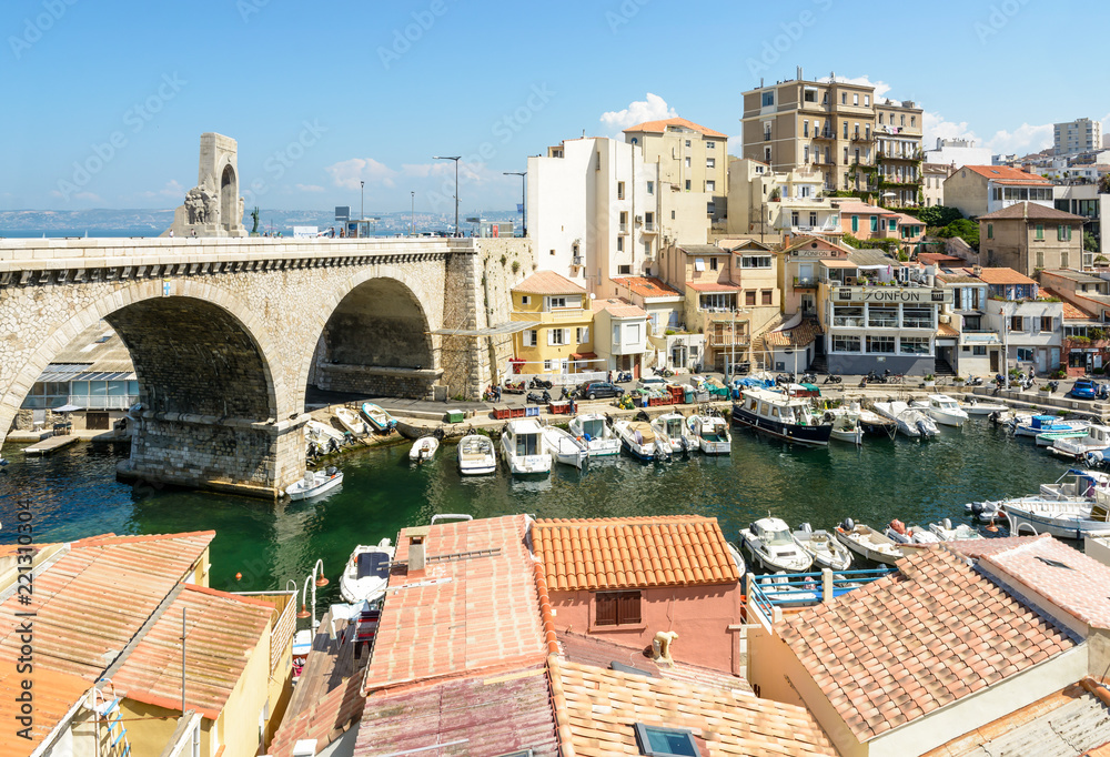 View from above of the small fishing port of the Vallon des Auffes in Marseille, France, with traditional and modern boats, colorful cabanons, the bridge of the Kennedy corniche and the Orient gate.