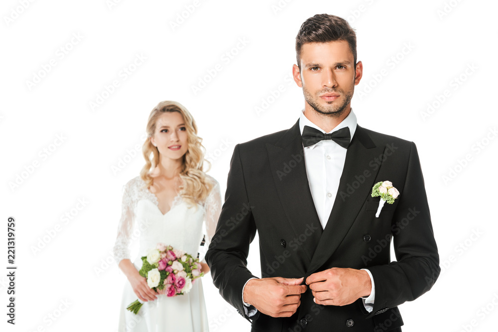 handsome young groom buttoning jacket and looking at camera with bride standing blurred on background isolated on white handsome young groom buttoning jacket and looking at camera with bride standing blurred on background isolated on white