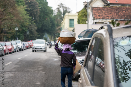 Vendor with basket of bread on head on Hanoi street, Vietnam. Two rows of cars parking on street. Concept of contrast between living level