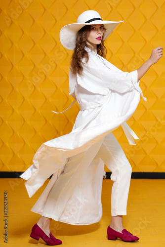 Full-length studio fashion portrait of young beautiful model wearing long white trench coat, trousers, wide-brimmed hat, loafers, posing on yellow background