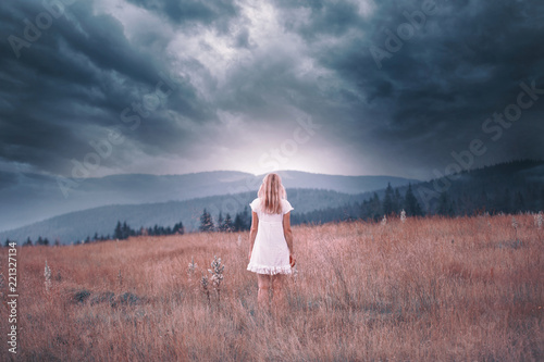 Back view of a blonde woman in white dress standing on the top of the hill with dark stormy sky background. Color tone filter effect used. photo