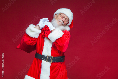 Christmas. Kind and tired Santa Claus in white gloves carries a red bag with gifts over his shoulder. Isolated on red background.