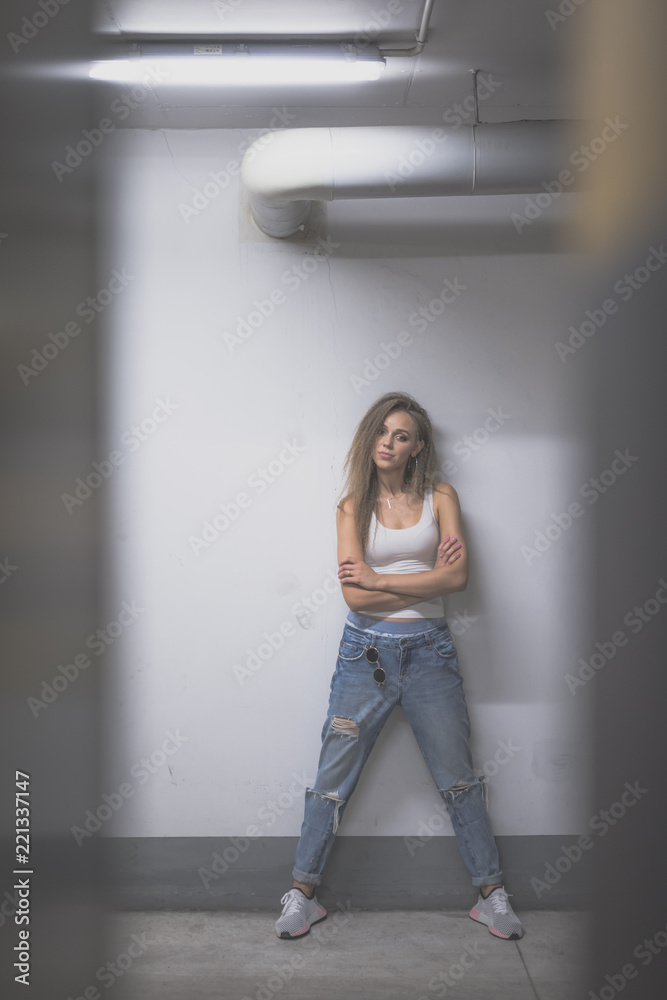 Portrait of young beautiful woman wearing white tank shirt and blue jeans with long hair at underground garage Portrait of young beautiful woman wearing white tank shirt and blue jeans with long hair at underground garage