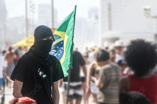 anarchist black-bloc man and a brazilian flag photo